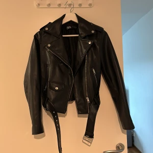 Svart skinnjacka från ZARA med bälte - Säljer en svart skinnjacka från ZARA i bikerstil. Jackan har silvriga dragkedjor, knappar och ett bälte i midjan. Klassisk krage och flera fickor framtill. Perfekt för dig som vill ha en edgy look.