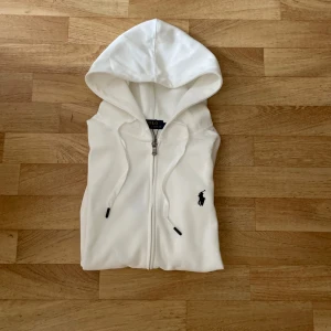 Helt ny vit zip hoodie från Ralph Lauren - Säljer en stilren vit zip hoodie från Ralph Lauren med dragkedja. Hoodien har en svart broderad logotyp på bröstet och är perfekt för lager-på-lager-stil.