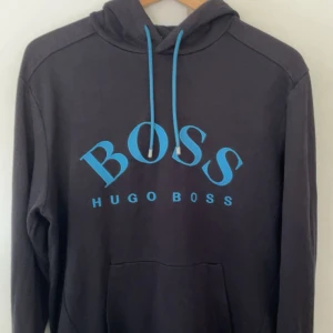 Svart Hugo Boss med blå logga - Säljer en svart hoodie från Hugo Boss med stor blå BOSS-logga framtill. Tröjan har huva med dragsko och en klassisk känguruficka. Ribbstickade muddar vid ärmslut och nederkant. Perfekt för en avslappnad stil. Längst ut på vänstra armen har hoddien en ficka med dragkedja, perfekt om du vil gömma något litet. Du ser fickan på bild 3. Hoddien är knappast andvänd.                               Det ända problemet är att den har ett pyttelite hål vid fickan längst ut på vänster arm. Men inget som syns
