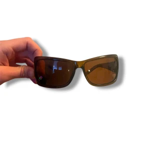 Bruna solglasögon med bred båge - Snygga solglasögon från Aolise med bred, brun båge och mörka linser. Bågarna har en diskret randig detalj i grönt, vitt och rött på sidan. Perfekta för soliga dagar och ger en cool look.