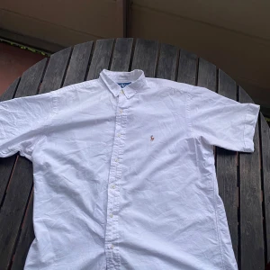 Vit kortärmad skjorta från Ralph Lauren - Klassisk vit kortärmad skjorta från Ralph Lauren med broderad logga på bröstet. Skjortan är tillverkad i 100% bomull och har knappar framtill. Perfekt för en stilren och avslappnad look. Nytvättad, vi stryker den åt dig innan vi fraktar. Skjortan har en millimeter stor öppning vid högra armhålan, som ni kan se på sista bilden är den minimal och syns knappt.