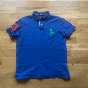 Ralph lauren piké - Säljer en blå piké t-shirt från Ralph Lauren med klassisk krage och två knappar. Stor grön broderad logga på bröstet och en orange siffra 3 på ärmen. Perfekt för en sportig och stilren look.