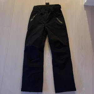 Svarta täckbyxor från VOLC - Svarta täckbyxor från VOLC med dragkedjor vid fickorna och justerbar midja. Perfekta för kalla dagar och vinteraktiviteter. Byxorna har raka ben och är tillverkade i ett slitstarkt material som står emot väder och vind.