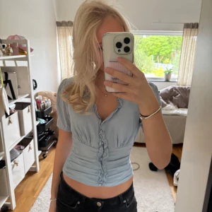 Ljusblå topp med volangdetaljer - Säljer en jättefin ljusblå topp från Guess med korta ärmar och volang längs knäppningen. Toppen har v-ringning och är perfekt till sommaren.💕