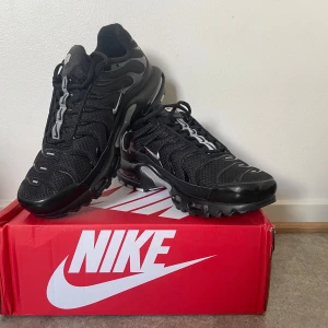 Nike Air Max Plus svarta sneakers - Säljer ett par svarta Nike Air Max Plus sneakers med silvriga detaljer och synlig Air Max-dämpning i sulan. Skorna har snörning och en sportig design med mesh och syntetmaterial. Perfekta för dig som gillar streetwear och komfort.