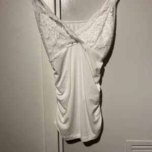 Vit spetsdetaljerad linnetopp - Säljer en vit linnetopp med vackra spetsdetaljer upptill och liten rosett framtill. Toppen har smala axelband och ärmlös design, perfekt för varma dagar eller som lager under andra plagg.