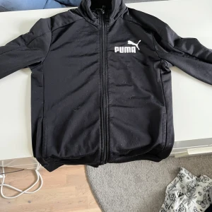 Svart träningsjacka från Puma - Svart träningsjacka från Puma med vit logga på bröstet och dragkedja framtill. Jackan har hög krage och långa ärmar. Perfekt för sportiga tillfällen och enkel att matcha med andra plagg.