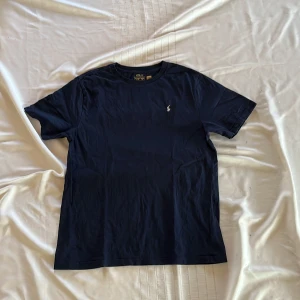 Mörkblå t-shirt från Polo Ralph Lauren - Säljer en mörkblå t-shirt från Polo Ralph Lauren med klassisk logga broderad på bröstet och stor POLO-tryck på ryggen. T-shirten har rund hals och är kortärmad. Perfekt för en stilren och avslappnad look. Går att pruta och såklart äkta.