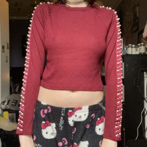 Vinröd croppad tröja med pärlor - Trendig vinröd croppad tröja med ribbad struktur och pärldetaljer längs ärmarna. Långärmad modell med rund halsringning, perfekt för att lyfta din outfit.