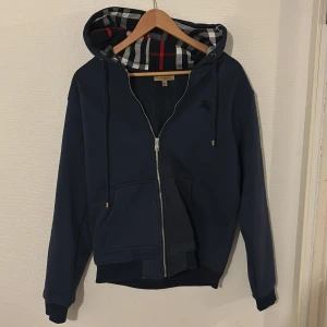 Marinblå hoodie från Burberry - Säljer en marinblå hoodie från Burberry med dragkedja och klassisk rutig insida på huvan. Jackan har broderad logga på bröstet, två fickor framtill och justerbar huva med snörning. Perfekt för dig som gillar stilrena detaljer och premiumkänsla. Original storleken är XL men hade sagt typ en M/L