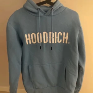 Blå HOODRICH hoodie med tryck - Säljer en blå hoodie från HOODRICH med vit logga framtill. Tröjan har huva med snörning och en stor magficka. Mjuk och skön, perfekt för chill dagar. Klassisk streetwear-stil med avslappnad passform.