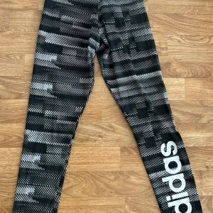 Svarta Adidas leggings med mönster XS - Snygga svarta leggings från Adidas i storlek XS med vit logga längs benet. De har ett grafiskt mönster i vitt och grått och är tillverkade i stretchigt material som passar perfekt till träning eller chill. Tight passform och hög midja för en sportig look.