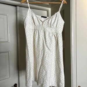 Vit blommig kortklänning med knyt - Supersöt vit kortklänning från Brandy Melville med tunna axelband, små blommor och knyt i ryggen. Klänningen har v-ringning, knappar framtill och är perfekt för varma dagar. Klänningen är i jättebra skick och har bara användts en gång. 