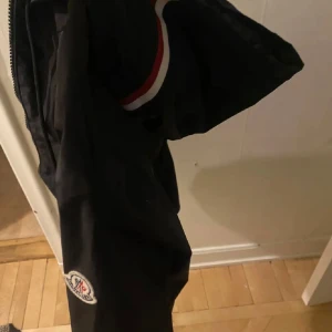 Svart vindjacka från Moncler med huva - Svart vindjacka från Moncler med huva och dragkedja framtill. Jackan har Moncler-logga på ärmen och snygga rödvita detaljer på huvan. Perfekt för dig som vill ha en stilren och sportig look. Två fickor med knappar framtill. Pris kan diskuteras. NFC funkar oxå 
