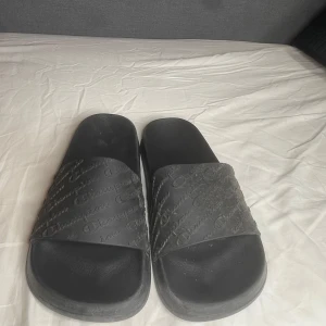 Svarta slides från Champion - Svarta slides från Champion med präglad logga över hela remmen. Skorna har en platt sula och öppen tå, tillverkade i syntetmaterial. Perfekta för chill dagar eller att slänga på sig efter träningen. Jag gör med på att förhandla om biligarr pris!!! (De är inte lika smutsiga som de ser ut)