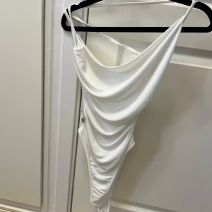 Vit body med snörning - Supersnygg vit body med draperad framsida och tunna axelband. Bodyn har omlottkänsla och snörningsdetaljer på sidan för en cool och trendig look. Materialet är glansigt och känns lyxigt mot huden, perfekt för att sticka ut.