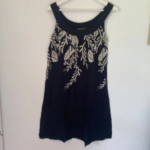 Svart kortklänning Twist & Tango - Svart kortklänning från Twist & Tango med breda axelband och beige bladmönster upptill. Klänningen har en luftig passform och är ärmlös, perfekt för varma dagar. Snygg och enkel att styla med sneakers eller sandaler.