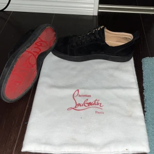 Svarta Christian Louboutins - Snygga svarta sneakers från Christian Louboutin i mocka med klassisk röd sula och rund tå. Skorna har svarta snören och diskreta detaljer, samt en lyxig känsla med beige innersida. Perfekta för dig som vill sticka ut med exklusiv stil.