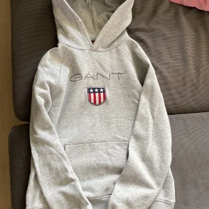 Grå hoodie från GANT med brodyr - Snygg grå hoodie från GANT med broderad logga och sköld på bröstet. Klassisk känguruficka framtill och stor huva. Mjuk bomullsblandning och långa ärmar. Perfekt för en chill och stilren look.