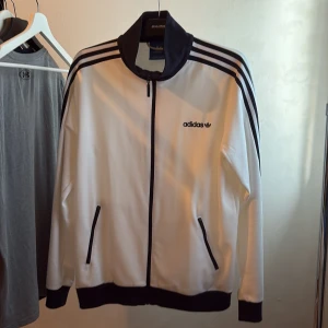 Vit Adidas track jacket med svarta detaljer - Säljer en klassisk vit Adidas track jacket med svarta ränder på axlarna och svarta muddar. Jackan har dragkedja framtill, två fickor och Adidas-logga på bröstet. Perfekt för en sportig och stilren look.