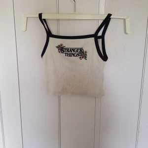 Stranger Things linne från H&M - Vit ribbad linne-topp från H&M x Netflix med svarta kanter och Stranger Things-brodyr framtill med blommiga detaljer. Smala axelband och croppad passform, perfekt för dig som älskar serien och vill ha en cool och bekväm look.