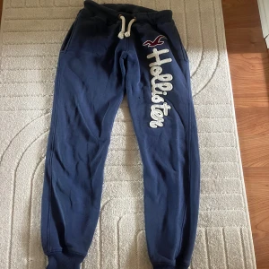 Lågmidjade mjukisbyxor från Hollister - Mörkblå mjukisbyxor från Hollister med vit snörning i midjan och stor broderad logga längs ena benet. Byxorna har resår i midjan och vid bensluten för en skön passform. Perfekta för chill eller träning.