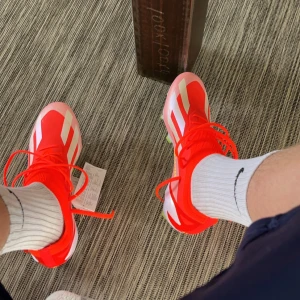 Röda adidas fotbollsskor med vita detaljer - Säljer ett par riktigt snygga fotbollsskor från adidas i en skarp röd färg med vita ränder och detaljer. Skorna har snörning och en modern, sportig design som sticker ut på planen. Perfekta för dig som vill ha stil och funktion på matchen.