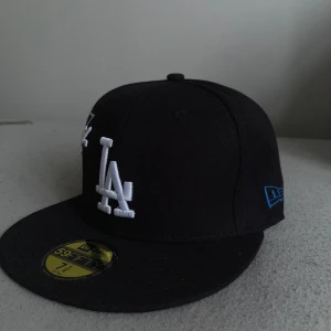 LA Dodgers svart keps New Era 59FIFTY - Svart LA Dodgers keps från New Era 59FIFTY med broderad vit LA-logga och Dodgers-märke på framsidan. Platt skärm med grå undersida och blå detalj på sidan. Klassisk basebollstil, perfekt för dig som gillar streetwear.