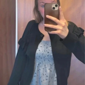 Svart kofta med volangdetaljer - Svart kofta med rund hals och volangdetaljer på axlarna. Långärmad och stängs med en knapp upptill. Enkel och stilren modell som passar perfekt till lager-på-lager. Mjuk och lätt materialkänsla.