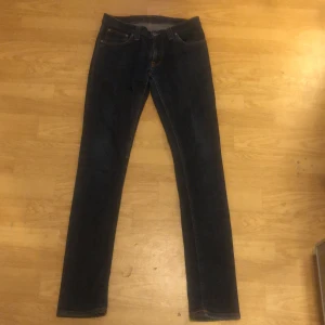 Mörkblå slim jeans från Nudie Jeans - Snygga mörkblå skinny jeans från Nudie Jeans med klassiska orange sömmar och broderade bakfickor. Jeansen har fem fickor, normal midja och är tillverkade i slitstark denim. Perfekta för dig som gillar en smal passform och stilren look. Storleken är 28 men passar som 29/30. Jeansen är i nyskick, inte använda fler än 5 ggr.