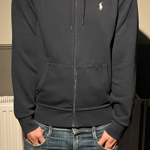 Mörkblå hoodie från Ralph Lauren - Mörkblå hoodie från Ralph Lauren med dragkedja, huva och snörning. Klassisk logga broderad på bröstet och känguruficka framtill. Skön passform och perfekt för chill dagar. Materialet känns mjukt och är troligtvis bomull eller bomullsblandning. Äkta såklart!