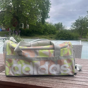 Adidas träningsväska med pastellmönster - Säljer en Adidas träningsväska med stort logotryck och coolt pastellfärgat mönster i beige, limegrönt och grått. Väskan har rejäla handtag, dragkedja och meshficka på sidan. Perfekt storlek för gymmet eller sportaktiviteter.Har lite fläckar men inget man märker av ordinarie pris är 367 kr.
