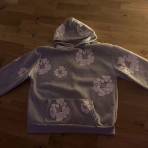 Grå hoodie med blommor från Denim Tears - Säljer en grå hoodie från Denim Tears med stora ljuslila blommor tryckta över hela tröjan. Tröjan har huva, magficka och långa ärmar. Perfekt för dig som vill sticka ut med ett unikt blommönster och streetwear-vibe.