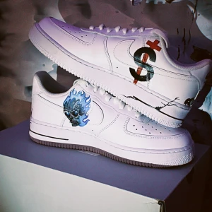 Nike Air Force 1  - Unika vita Nike Air Force 1 sneakers med handmålade motiv som blå flammande dödskalle, röda och svarta symboler samt coola tecknade ansikten. Skorna har klassisk låg siluett, snörning och platt sula. Perfekta för dig som vill sticka ut med streetstyle och konstnärlig vibe.