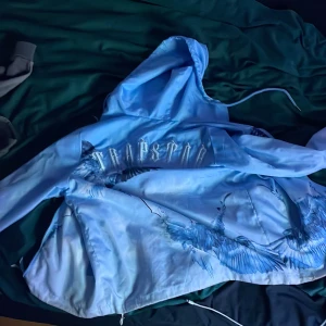 Blå vindjacka Trapstar med fågeltryck - Säljer en blå vindjacka från Trapstar med snyggt fågeltryck och logga på ryggen. Jackan har huva, dragkedja framtill och dragsko nertill för extra passform. Perfekt för dig som vill sticka ut med streetstyle-vibe. Storlek S men passar xs kort vid armarna och midjan 