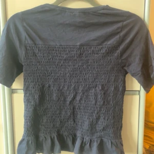 Mörkblå topp med volang från Gina Tricot - Supersöt mörkblå topp från Gina Tricot med smockad front och volangkant nertill. T-shirten har korta ärmar och rund halsringning. Perfekt för dig som gillar en enkel men ändå trendig look.