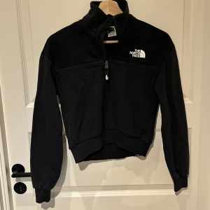 Svart fleecejacka från The North Face XS - Svart fleecejacka från The North Face i storlek XS. Jackan har hög krage, halv dragkedja och broderad logga på bröstet. Mjuk och varm med ribbade muddar vid ärmslut och nederkant. Perfekt för kyliga dagar och enkel att matcha med allt.