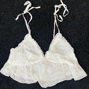 Vit broderad linnetopp med knytband - Superfin vit linnetopp med broderade hål och volangkant. Toppen har smala axelband som knyts och en lätt croppad passform. Perfekt för varma dagar, söt med sina detaljer.