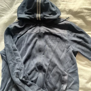 Mörkblå hoodie från Hugo Boss - Snygg mörkblå hoodiejacka från Hugo Boss med dragkedja hela vägen, huva med snörning och diskret broderad logga på bröstet. Jackan är i mjuk bomull och har ribbade muddar vid ärmsluten. Perfekt för en chill och stilren look.