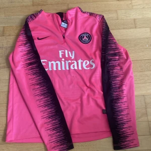PSG långärmad rosa Nike fotbollströja - Paris Saint-Germain långärmad fotbollströja i stark rosa med svarta detaljer och grafiskt mönster längs ärmarna. Tröjan har halv dragkedja, broderad Nike-logga och PSG-emblem på bröstet. Materialet är lätt och andas, perfekt för träning eller match.