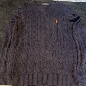 Svart kabelstickad tröja Polo Ralph Lauren - Kabelstickad svart tröja från Polo Ralph Lauren med klassisk orange logga broderad på bröstet. Tröjan har rund halsringning, ribbade muddar och är långärmad. Perfekt för dig som gillar stilren och tidlös design.