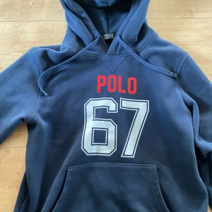 Blå hoodie Polo Ralph Lauren L - Mörkblå hoodie från Polo Ralph Lauren med stor vit 67-print och röd POLO-text på bröstet. Klassisk huva med snörning och känguruficka framtill. Perfekt för dig som gillar sportig och avslappnad stil. Storlek L men passar som M. 
