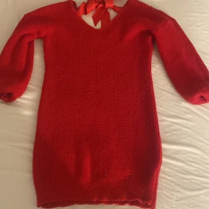 Röd stickad kortklänning med rosett - Superfin röd stickad kortklänning med lång ärm och v-ringning. Snygg detalj med bred röd rosett i nacken som knyts. Perfekt för dig som vill ha en mysig men ändå stilren look.