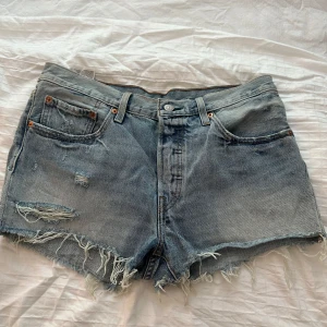 Levi's ljusblå jeansshorts med slitningar - Ljusblå jeansshorts från Levi's med råa, fransiga kanter och slitna detaljer framtill. Klassisk femficksmodell med knapp och dragkedja. Perfekta för sommaren och ger en avslappnad vibe. Ikonisk läderpatch bak och tydlig Levi's-märkning.