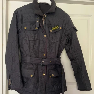 Barbour jacka - Snygg svart quiltad jacka från Barbour med bälte i midjan, guldiga knappar och dragkedja. Jackan har flera fickor, hög krage och klassisk Barbour-logga på bröstet. Perfekt för dig som gillar stilren och tidlös design.