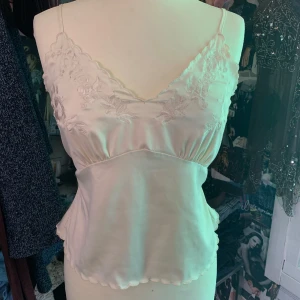 Vit sidenlinnetopp från ZARA - Supersöt vit linnetopp från Zara i siden med tunna axelband och broderade blommor upptill. Toppen har v-ringning, lätt figursydd passform och vågig kant nertill. 