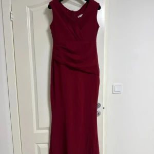 Vinröd långklänning med omlott - Snygg vinröd långklänning med omlott-detalj upptill och drapering i midjan. Ärmlös modell med v-ringning och dragkedja bak. Perfekt för dig som vill sticka ut med en elegant och stilren look.