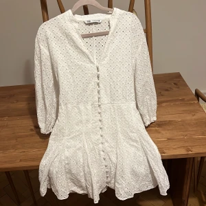 Vit kort klänning med spets - Superfin vit kort klänning från Zara med broderad spets, knappar framtill och knytband i ryggen. Klänningen har trekvartsärmar med resår och ett sött blommigt hålmönster i bomull. Perfekt för sommarens alla tillfällen!