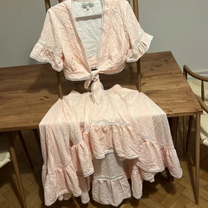 Ljusrosa rutig långkjols set By Malina - Superfin långkjol med knytblus från By Malina i ljusrosa och vitt rutigt bomullstyg. Kjolen har volanger, blusen knyt framtill, korta ärmar med volangkant och fina spetsdetaljer. Perfekt för sommarens alla tillfällen! Obs se olika storlekar