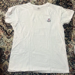 Vit t-shirt  - Vit basic t-shirt från B&C Collection i storlek S, med rund hals och korta ärmar. På bröstet sitter en broderad Moncler-inspirerad patch som ger en clean och stilren vibe. Perfekt till jeans eller shorts för en enkel look.